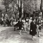 Fotografia przedstawia ludzi idących w pochodzie: mężczyźni trzymają flagi, elegancko ubrane kobiety prowadzą dzieci.