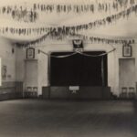 Fotografia przedstawia pomieszczenie: z przodu scena, bo bokach pod ścianami rzędy krzeseł. Z sufitu zwisają wstążki. Nad sceną orzeł, po bokach dwa portrety. Na scenie napis „Witajcie”.