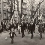 Fotografia przedstawia mężczyzn w garniturach kroczących z flagami. Przed grupą maszeruje mały chłopiec w dwukolorowej chustce na szyi.