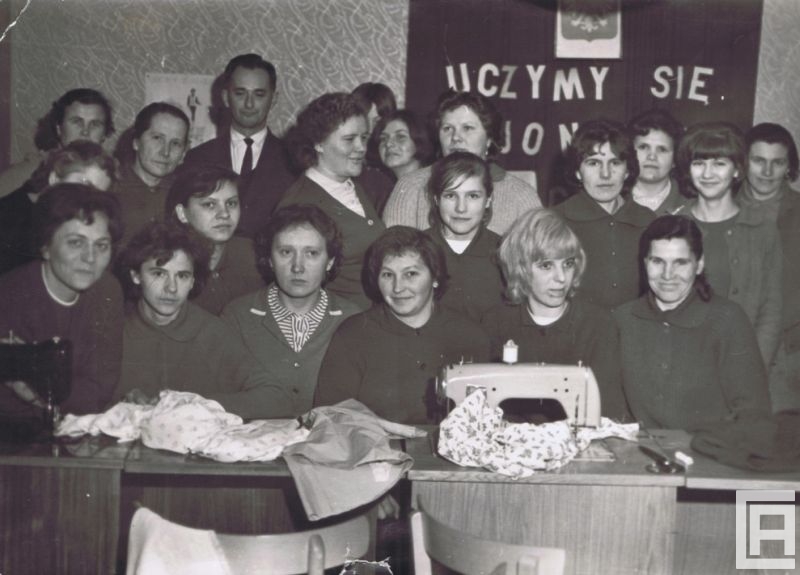 Fotografia przedstawia grupę kobiet i mężczyznę zebranych przed stolikami, na których złożono materiały oraz maszynę do szycia. Z tyłu godło oraz fragment napisu "Uczymy się".