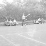 Fotografia przedstawia osobę zamachującą się rakietą tenisową podczas gry. Z tyłu na dwóch ławkach siedzi pięć osób.