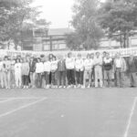 Fotografia przedstawia grupę osób pozującą do zdjęcia na korcie tenisowym. Na płocie napis "Witamy uczestników Spartakiady".