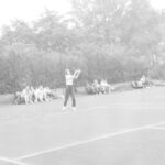 Fotografia przedstawia kobietę, która właśnie odbiła piłkę tenisową. Z tyłu na ławkach widoczne liczne grono przypatrujących się grze.