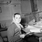 Fotografia przedstawia siedzącą kobietę, która nożyczkami przycina rysunki na płachtach papieru.