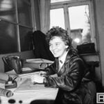 Fotografia przedstawia kobietę siedzącą przy biurku nad rysunkiem technicznym. W ręku ma piórko, obok widoczny pojemnik z tuszem kreślarskim.
