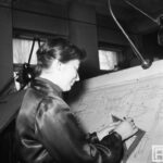 Fotografia przedstawia kobietę przy tablicy kreślarskiej - zaznacza coś na rysunku technicznym przy pomocy ołówka i drewnianej ekierki.