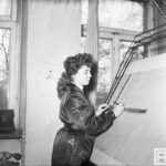 Fotografia przedstawia kobietę przy tablicy kreślarskiej - zaznacza coś na rysunku technicznym przy pomocy ołówka i metalowej ekierki.