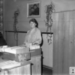 Fotografia przedstawia kobietę przeglądającą dokumenty. Nad nią widoczna reprodukcja obrazu "Śpiący Mietek" Stanisława Wyspiańskiego.
