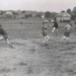 Fotografia przedstawia grupę osób w strojach sportowych z numerami na plecach. W tle widoczna grupa kibiców oraz nieliczne zabudowania.