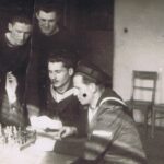 Fotografia przedstawia pięciu mężczyzn w strojach marynarskich. Dwóch z nich gra w szachy, pozostali przyglądają się.
