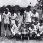 Fotografia przedstawia grupę mężczyzn pozujących do zdjęcia w strojach sportowych. Na środku, na krześle, widoczny puchar. W tle widoczna piłka do gry wyrzucona w powietrze.