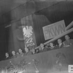 Fotografia przedstawia grupę osób ubranych uroczyście, w garniturach, mundurach wojskowych, siedzących w rzędzie. Za nimi orzeł bez korony z napisem "22 lipca" oraz tablica "XXI rocznica PKWN".