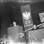 Fotografia przedstawia mężczyznę na mównicy z mikrofonem, obok gości w loży. Z tyłu widoczny orzeł bez korony i napis "22 lipca".