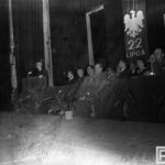 Fotografia przedstawia grupę osób ubranych uroczyście, w garniturach, mundurach wojskowych, siedzących w rzędzie. Za nimi orzeł bez korony z napisem "22 lipca" oraz tablica "XXI rocznica PKWN". Po lewej mężczyzna przemawia na mównicy do mikrofonu.