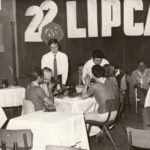Fotografia przedstawia ludzi siedzących przy stolikach. Na ścianie napis "22 lipca, 1944 1973 PKWN". Po lewej widoczny plakat "Zlot Młodych Przodowników Pracy i Nauki".