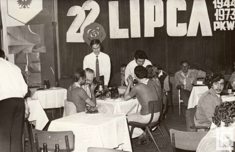 Fotografia przedstawia ludzi siedzących przy stolikach. Na ścianie napis "22 lipca, 1944 1973 PKWN". Po lewej widoczny plakat "Zlot Młodych Przodowników Pracy i Nauki".