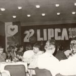 Audiodeskrypcja: Fotografia przedstawia ludzi siedzących przy stolikach. Na ścianie napis "22 lipca, 1944 1973 PKWN". Po lewej widoczny plakat "Zlot Młodych Przodowników Pracy i Nauki" oraz orzeł bez korony wpisany w kontury Polski.