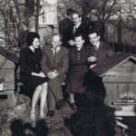 Fotografia przedstawia grupę mężczyzn i kobiet siedzących lub przechadzających się w pobliżu uli. Z tyłu widoczny dom.