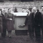 Fotografia przedstawia grupę kobiet i mężczyzn obok ula. W tle dom.