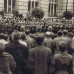 Fotografia przedstawia widok na tłum stojący wzdłuż ulicy. Po drugiej stronie żołnierze oddają honory.