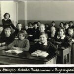 Fotografia przedstawia dwa rzędy ławek szkolnych z uczniami. Z lewej widoczna nauczycielka oraz fragment pieca kaflowego.
