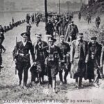 Fotografia przedstawia kolumnę polskich żołnierzy prowadzonych pomiędzy uzbrojonymi żołnierzami niemieckimi. Napis: Załoga Westerplatte w drodze do niewoli.