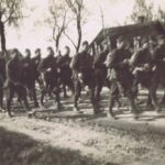 Fotografia przedstawia kolumnę uzbrojonych żołnierzy w mundurach maszerujących drogą. W tle drzewa i wiejskie zabudowania.