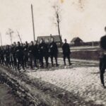 Fotografia przedstawia kolumnę uzbrojonych żołnierzy w mundurach maszerujących drogą. W tle drzewa i wiejskie zabudowania