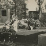 Fotografia przedstawia grób na cmentarzu, otoczony zapalonymi zniczami, świeżo przykryty wieńcami pogrzebowymi, nie ma jeszcze tablicy nagrobkowej.