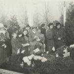 Fotografia przedstawia grupę osób – kobiet, mężczyzn i dzieci na cmentarzu. Grób, przy którym stoją, jest świeżo przykryty wieńcami pogrzebowymi oraz chryzantemami w donicach, nie ma jeszcze tablicy nagrobkowej.