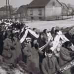 Fotografia przedstawia kolumnę ludzi odprowadzających trumnę podczas ceremonii pogrzebowej. Na niesionych wieńcach widoczne są wstęgi z napisami: „Wielebnemu Kanonikowi”, „Wielebnemu Księdzu”, „Księdzu Siostrzeńcowi”. W tle widoczne murowane domy.