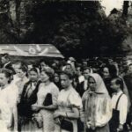 Fotografia przedstawia tłum ludzi odprowadzających trumnę podczas ceremonii pogrzebowej. W tle widoczne drewniane domy.