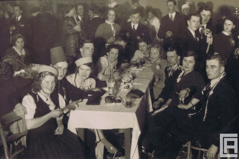 Fotografia przedstawia grupę ludzi na zabawie sylwestrowej. W centrum przy stoliku siedzą kobiety i mężczyźni. Widoczne karnawałowe nakrycia głowy, serpentyny.