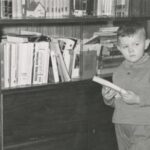 Fotografia przedstawia małego chłopca stojącego z książką przed domową biblioteczką.