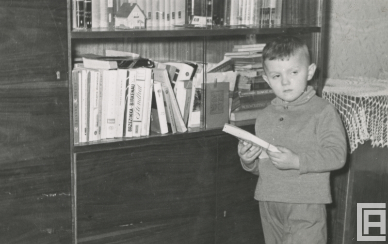 Fotografia przedstawia małego chłopca stojącego z książką przed domową biblioteczką.