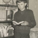 Fotografia przedstawia starszego chłopca stojącego z książką przed domową biblioteczką.