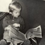 Fotografia przedstawia dziecko siedzące w głębokim fotelu, które przegląda dużą gazetę.