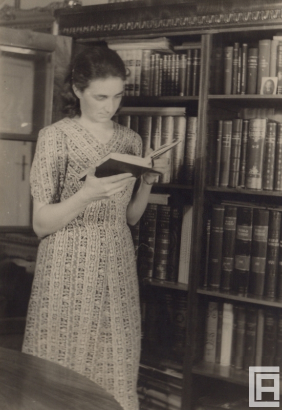 Fotografia przedstawia młodą kobietę stojącą przy witrynie domowej biblioteczki z książką w ręku.