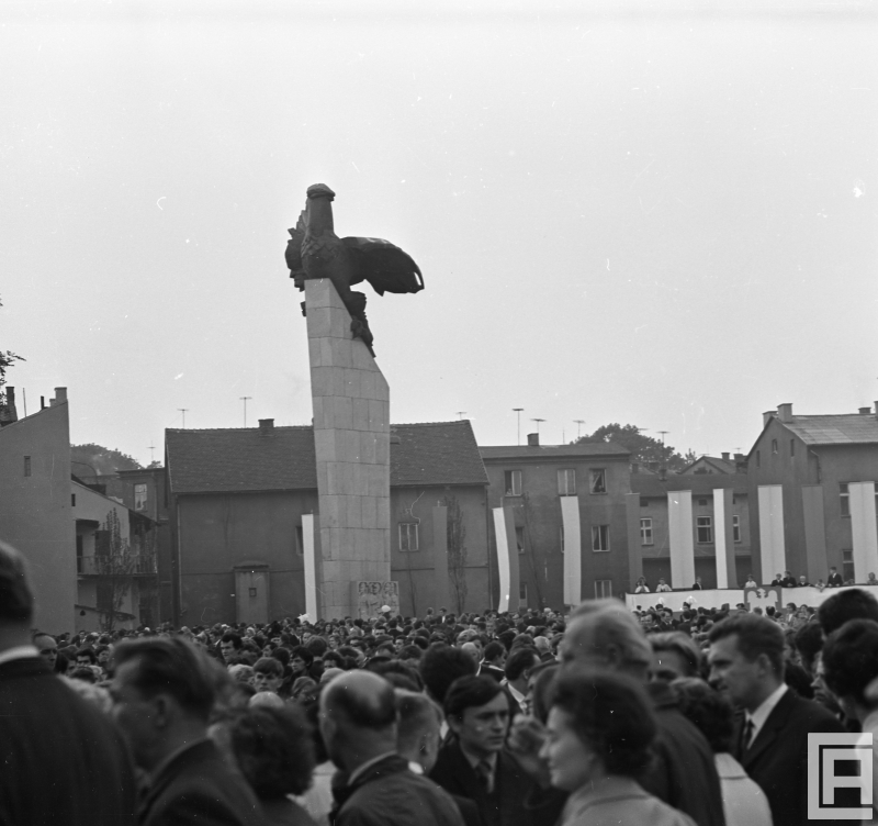Fotografia przedstawia profil pomnika orła na tle otoczenia placu. Widoczny tłum ludzi, wiszące flagi państwowe.