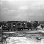 Fotografia przedstawia plac budowy naprzeciw bloku mieszkalnego. Z lewej strony widoczny częściowo cokół przyszłego pomnika z rusztowaniem. Z prawej samochód z naczepą.