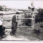 Fotografia przedstawia dwóch mężczyzn idących z lewej strony - pierwszy jest listonoszem, drugi ma na sobie garnitur. Po prawej stronie widocznych jest troje dzieci bawiących się przy drewnianym płocie - dwóch chłopców oraz dziewczynkę.