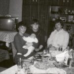 Fotografia przedstawia dwie kobiety i mężczyznę siedzących przy zastawionym stole. Jedna z kobiet trzyma niemowlę. Za nimi widoczny telewizor oraz meblościanka zawierająca serwis i fotografie.