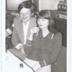 Fotografia przedstawia mężczyznę i kobietę siedzących przy stole i wznoszących toast. Za nimi widoczny jest telewizor.