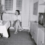 Fotografia przedstawia kobietę siedzącą na krześle. Po jej prawej stronie, na meblościance stoi telewizor.