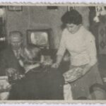 Fotografia przedstawia dwóch mężczyzn siedzących przy zastawionym stole, kobieta stojąca obok nakłada im jedzenie. W tle widoczny jest włączony telewizor.