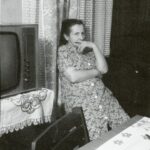 Fotografia przedstawia kobietę siedzącą pod oknem. Po jej lewej stronie – na nakrytym chustką stoliku stoi telewizor.