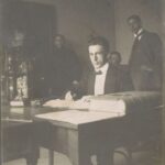 Fotografia przedstawia na pierwszym planie mężczyznę w garniturze siedzącego przy biurku. W tle widocznych jest trzech mężczyzn patrzących w obiektyw.