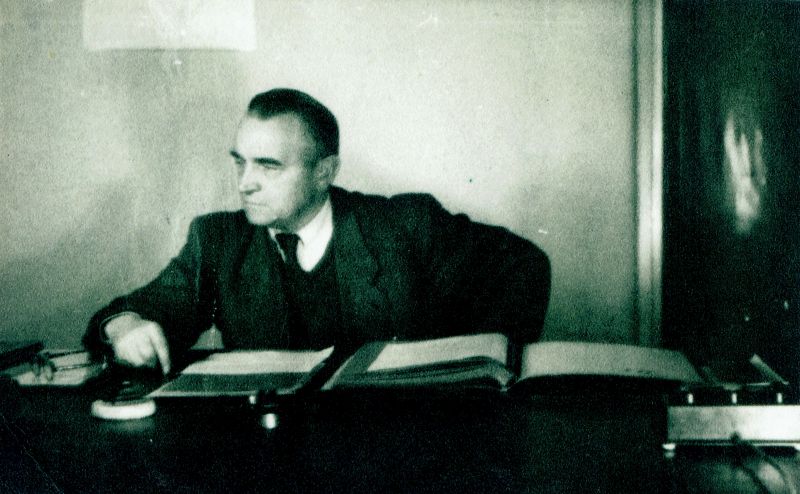 Fotografia przedstawia mężczyznę w średnim wieku w garniturze, z krawatem. Siedzi on za biurkiem, przed sobą ma rozłożone dokumenty.