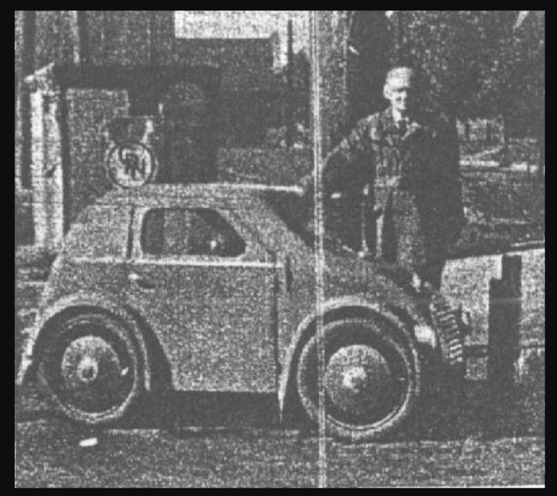 Czarno-biała fotografia. Małe auto osobowe, obok stoi pan w garniturze.
